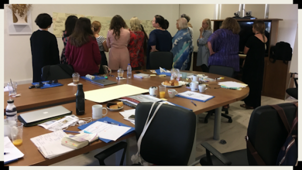 photo a group of people standing behind a conference table. They are looking at large pieces of paper on a wall, considering the words written on the paper.