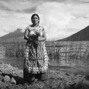 black and white photograph of Mayan woman standing against a landscape of mountains, water, tall grass, and clouds. She looks straight ahead, holding an handful of  flowers.