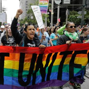 photo of large group of protesters carrying a rainbow banner with the word "unite"