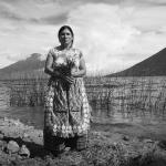 black and white photograph of Mayan woman standing against a landscape of mountains, water, tall grass, and clouds. She looks straight ahead, holding an handful of  flowers.