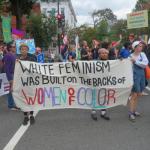 photograph of 3 young women (2 women of color and one white woman) holding a banner at a protest. the banner reads "White feminism was built on the backs of women of color"