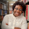 A smiling headshot of Dr. Moya Z Bailey, a black woman with glasses, leaning on a bookshelf